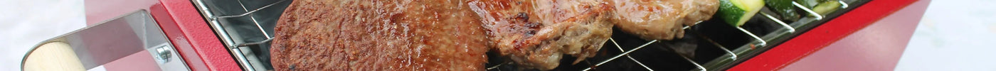 Barbecue portatile rosso con carne e zucchine sulla griglia in acciaio, fumo in uscita, ideale per esterni.