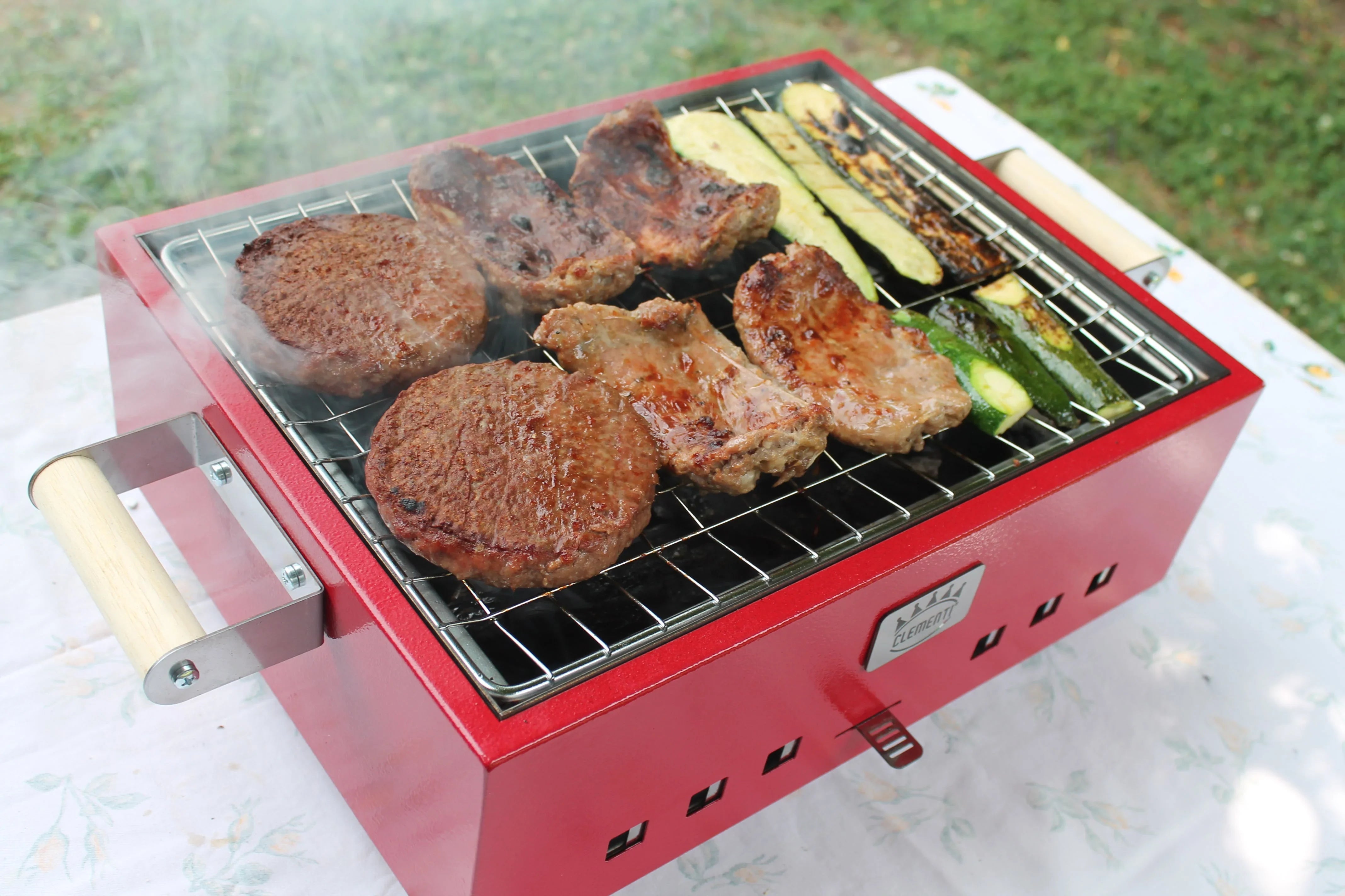 Barbecue portatile rosso con carne e zucchine sulla griglia in acciaio, fumo in uscita, ideale per esterni.