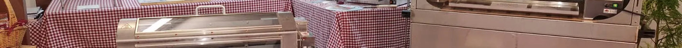 Barbecue e girarrosto in acciaio inox per cucina professionale e privata, esposti su tavoli con tovaglia a quadri
