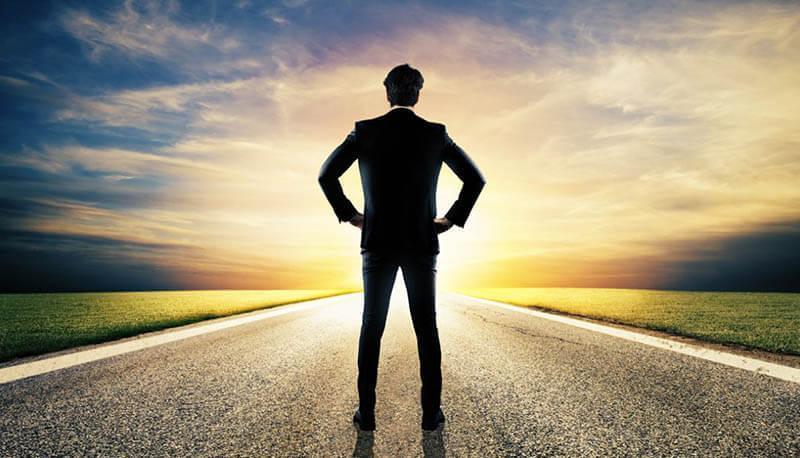 Uomo in abito che guarda un viale al tramonto, simbolo di successo e visione futura.